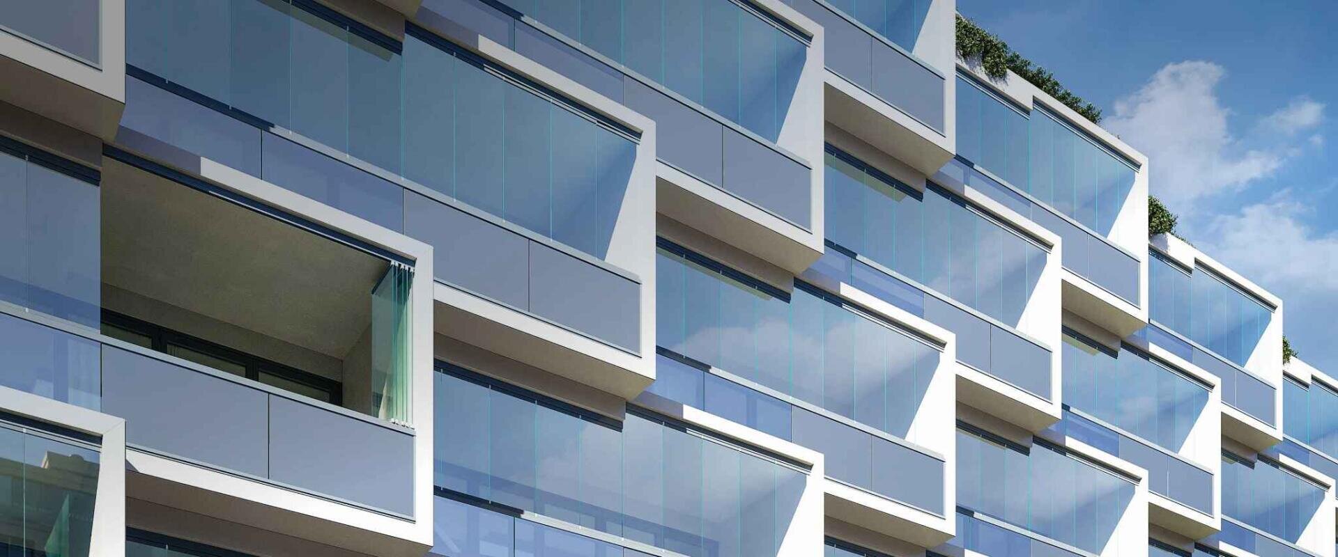 Moderne Gebäudefassade mit großen, eckigen Glasfenstern und weißen Rahmen, in denen sich der blaue Himmel und die Wolken spiegeln. Einige Fenster sind offen, so dass sich hinter den Glasscheiben Balkone befinden.