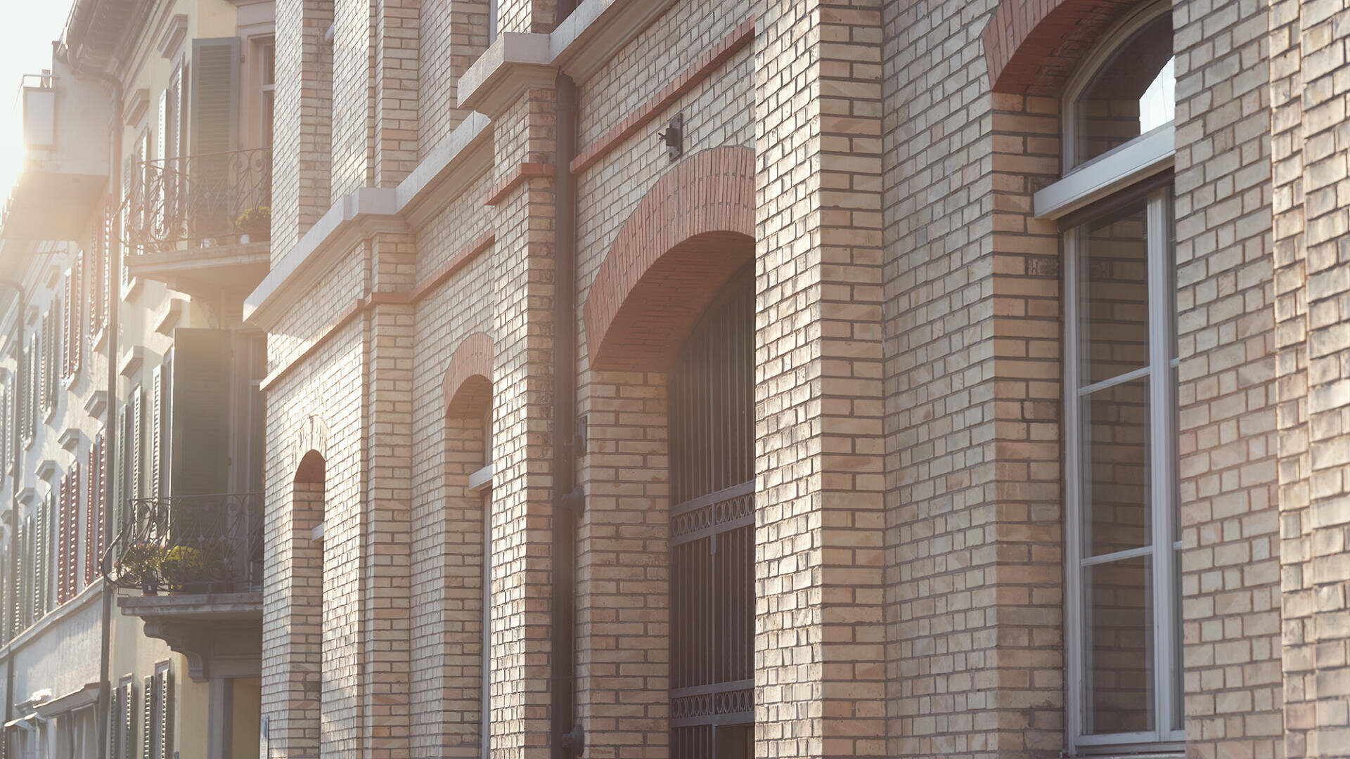 Sonnenbeschienene Nahaufnahme einer Backsteinfassade mit Bogenfenstern und dekorativem Mauerwerk, neben einem anderen Gebäude mit Balkonen und grünen Fensterläden.