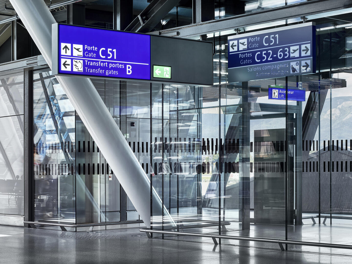 Ein modernes Flughafenterminal mit Glaswänden, reflektierendem Bodenbelag und großen blau-weißen Schildern, die auf die Flugsteige C51, C52-63 und die Transfer-Gates B hinweisen. Auch Richtungspfeile und Notausgangsschilder sind zu sehen.