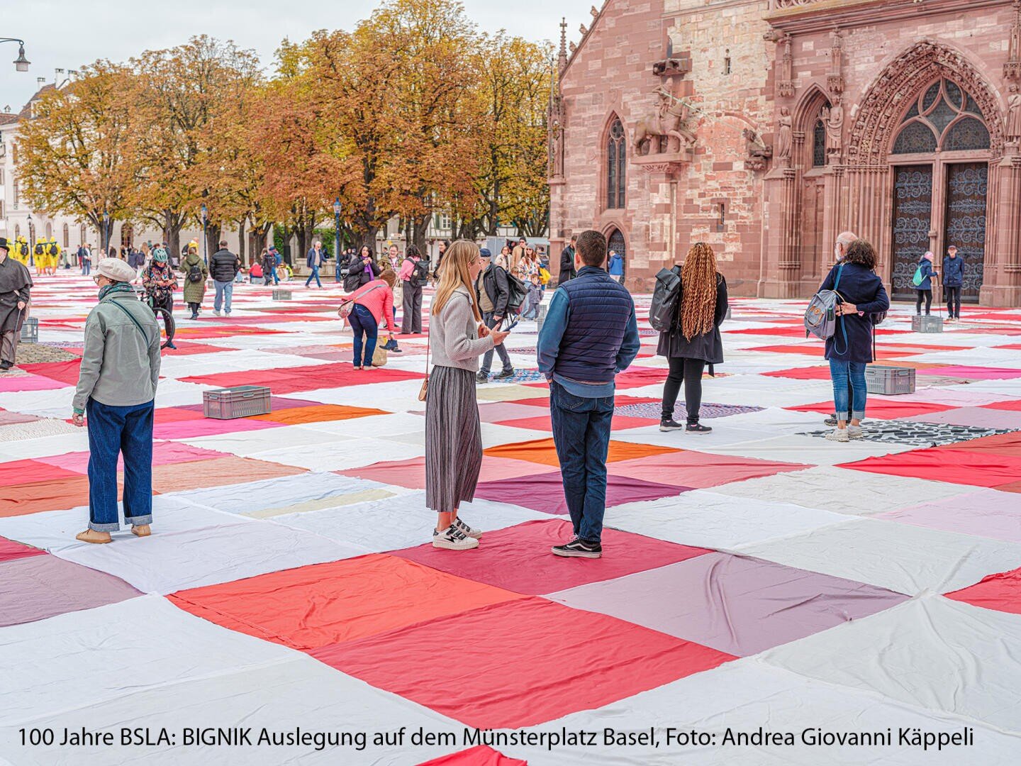 Die Menschen gehen und stehen auf einem großen Flickenteppich aus roten, rosafarbenen und weißen Stoffquadraten, der auf einem öffentlichen Platz in der Nähe einer historischen Kirche ausgelegt und von Bäumen umgeben ist.