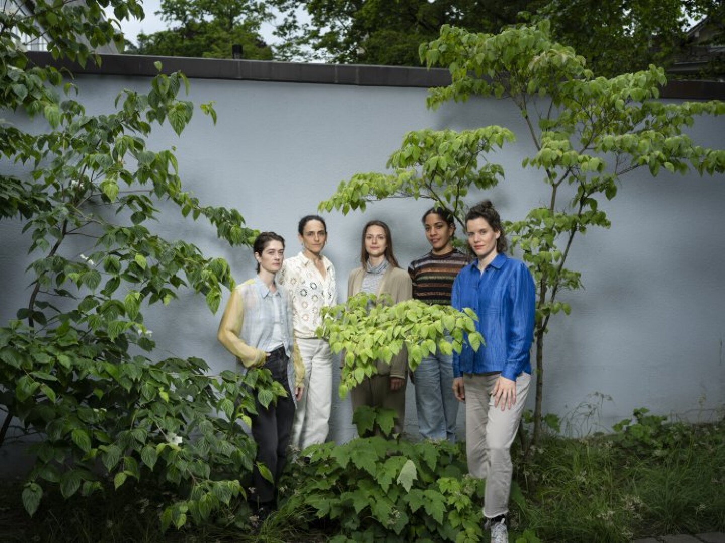 Fünf Personen stehen gemeinsam draußen vor einer hellblauen Wand, umgeben von grünen Pflanzen und Bäumen, und posieren für ein Gruppenfoto. Die Szene wirkt ruhig und natürlich.