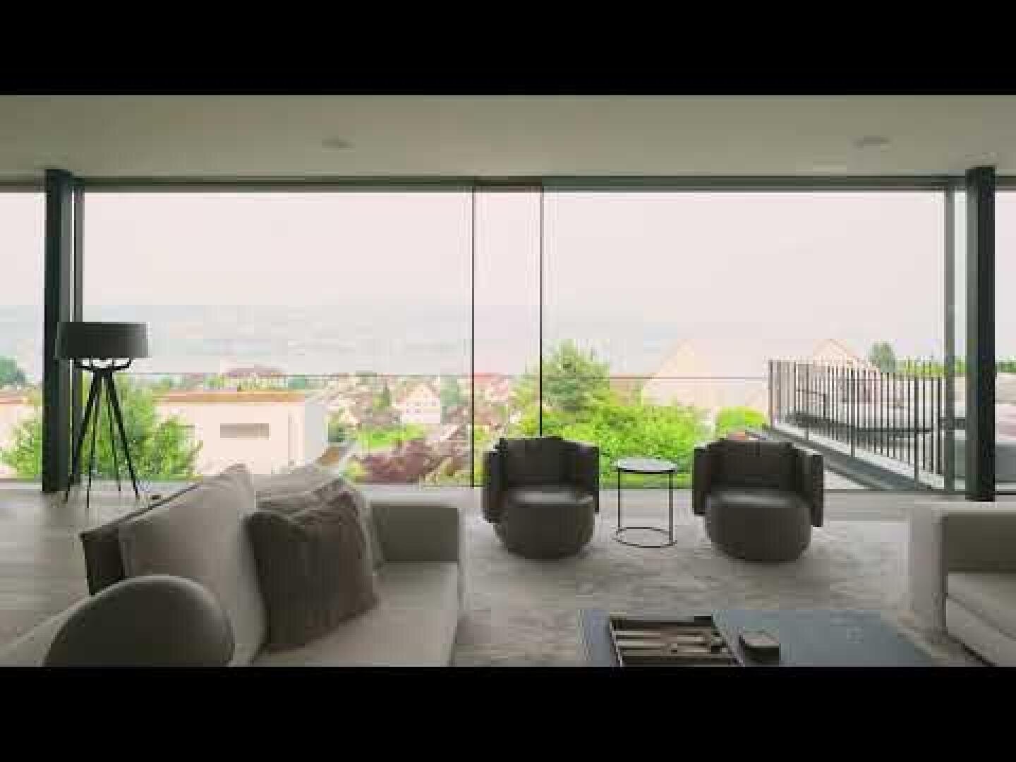 Ein modernes Wohnzimmer mit großen, raumhohen Fenstern, von denen aus man einen malerischen, dunstigen Blick auf Dächer, Grünflächen und entfernte Hügel hat. Der Raum verfügt über Sofas in neutralen Farbtönen, Sessel, eine Stehlampe und einen Balkon.