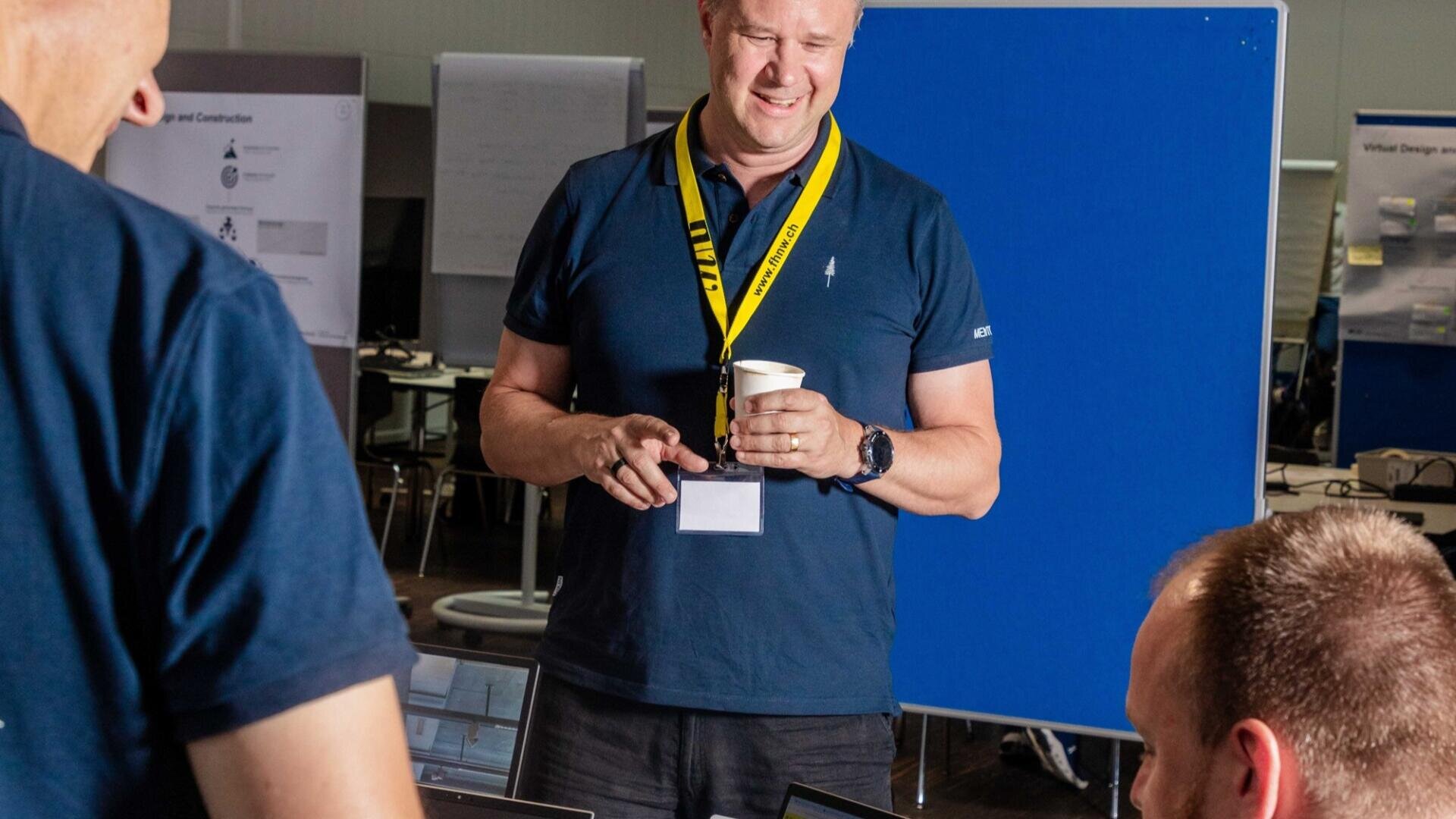 Ein Mann, der ein marineblaues Hemd, ein gelbes Schlüsselband und ein Namensschild trägt, lächelt und hält einen Pappbecher, während er steht und mit zwei sitzenden Kollegen in einer Büroumgebung mit Postern und blauer Trennwand hinter ihm spricht.