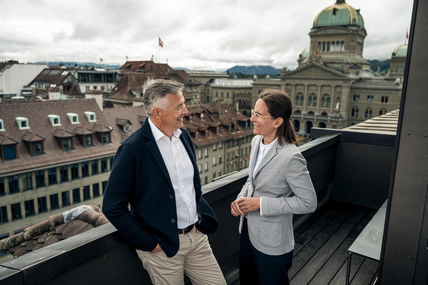 Zwei professionell gekleidete Erwachsene, ein Mann und eine Frau, stehen lächelnd auf einer Dachterrasse und unterhalten sich, während im Hintergrund historische Gebäude und ein Kuppelbau unter einem bewölkten Himmel zu sehen sind.
