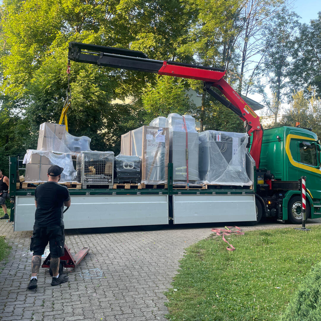 Ein Mann bedient einen Hubwagen neben einem großen grünen Lkw mit rotem Kran, der an einem sonnigen Tag mehrere verpackte und in Kisten verpackte Waren auf eine gepflasterte Fläche entlädt, die von Bäumen und Gras umgeben ist.
