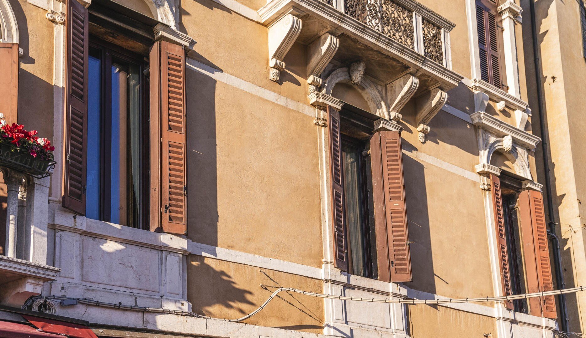 Gros plan sur une façade d'immeuble de couleur beige avec de hautes fenêtres, des volets bruns, une maçonnerie décorative et une jardinière de fleurs rouges sur un petit balcon ensoleillé.
