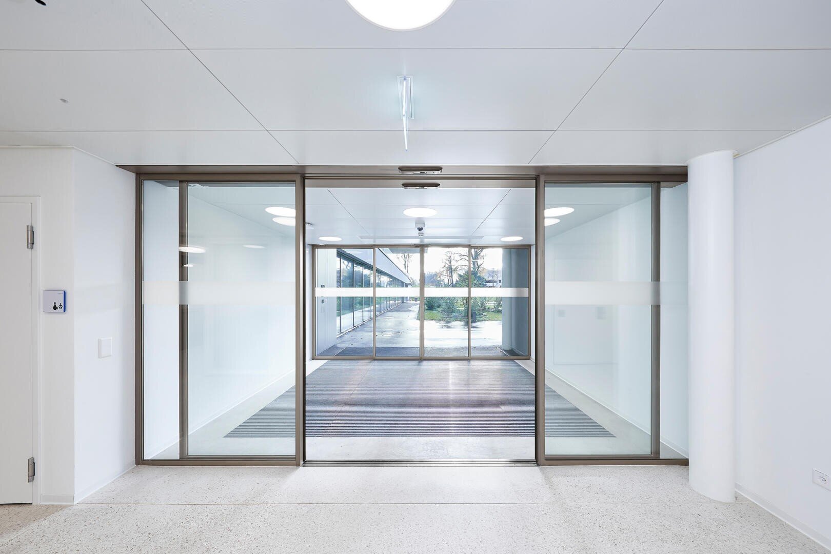 Ein moderner, heller Flur mit großen Glasschiebetüren und weißen Wänden, der zu einem Korridor mit Fenstern auf beiden Seiten und einem Blick ins Grüne führt.