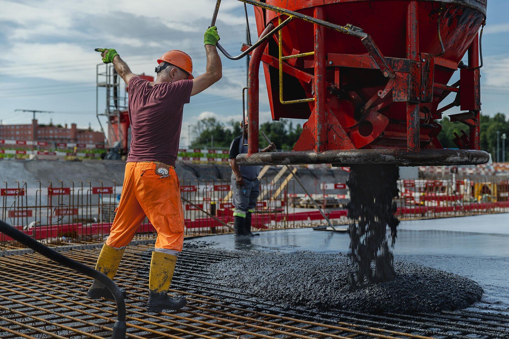 Ein Bauarbeiter in orangefarbener Hose und Schutzhelm steuert eine große rote Betongießmaschine, mit der er den nassen Beton auf ein Bewehrungsgerüst auf einer Baustelle leitet. Ein weiterer Arbeiter ist im Hintergrund zu sehen.