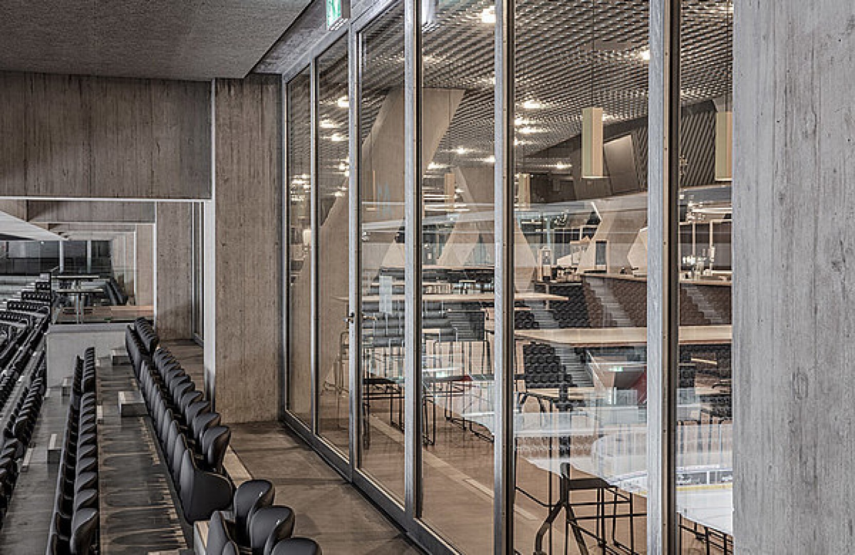 Reihen schwarzer Stühle stehen vor großen Glasfenstern mit Blick auf eine Indoor-Arena mit geometrischen Betonstrukturen, Metallgeländern und einer Decke mit Gittermuster.