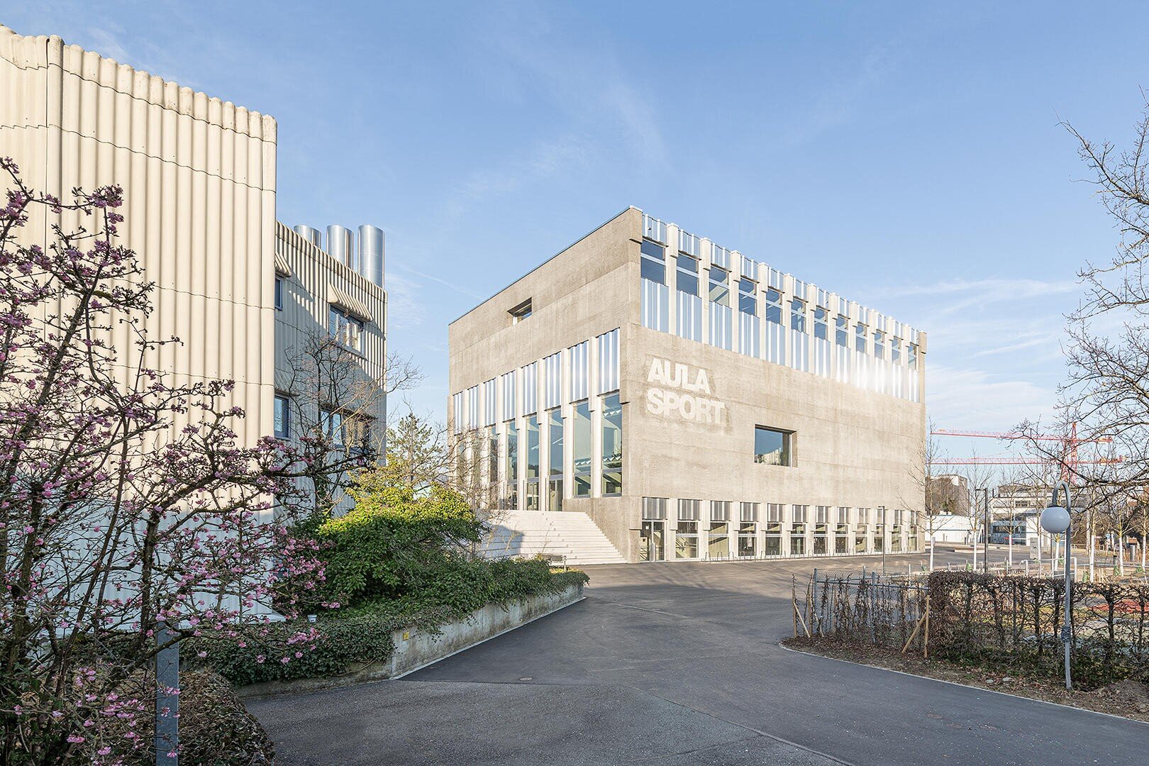 Ein modernes Betongebäude mit der Aufschrift AULA SPORT steht unter einem strahlend blauen Himmel, umgeben von Bäumen und Sträuchern, und ein gepflasterter Weg führt zum Eingang.