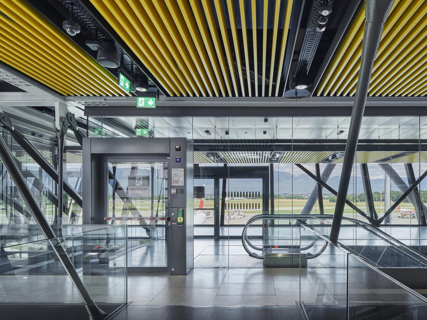 Intérieur d'un terminal d'aéroport moderne avec des murs en verre, des lattes de plafond jaunes, un ascenseur, un escalator et des panneaux de sortie de secours. La vue extérieure montre la piste d'atterrissage et les montagnes au loin.
