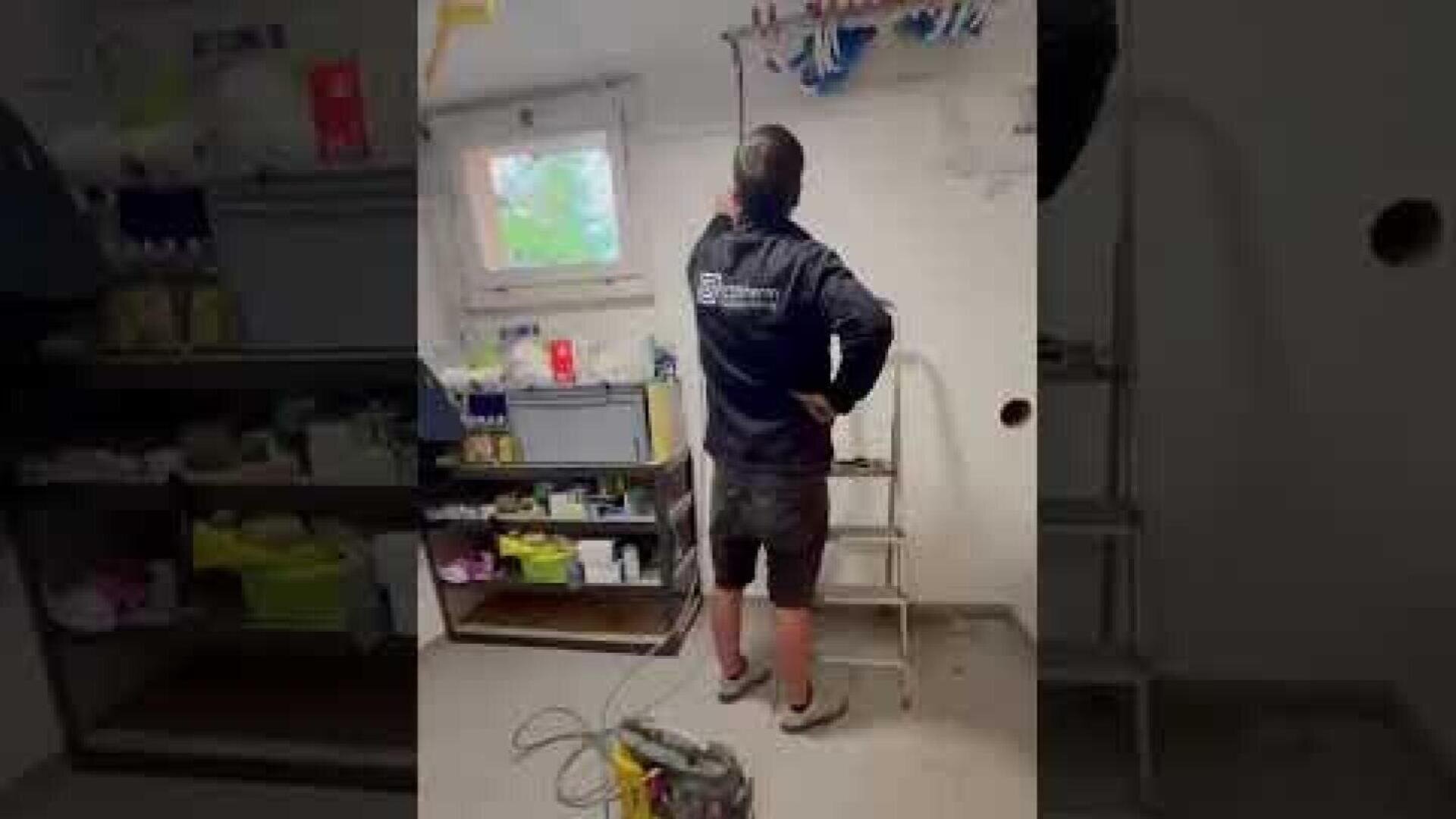 Eine Person in Arbeitskleidung steht auf dem Boden neben einer kleinen Stufenleiter in einem Hauswirtschaftsraum und benutzt ein Werkzeug. Im Hintergrund sind Regale mit verschiedenen Materialien und ein Fenster zu sehen.