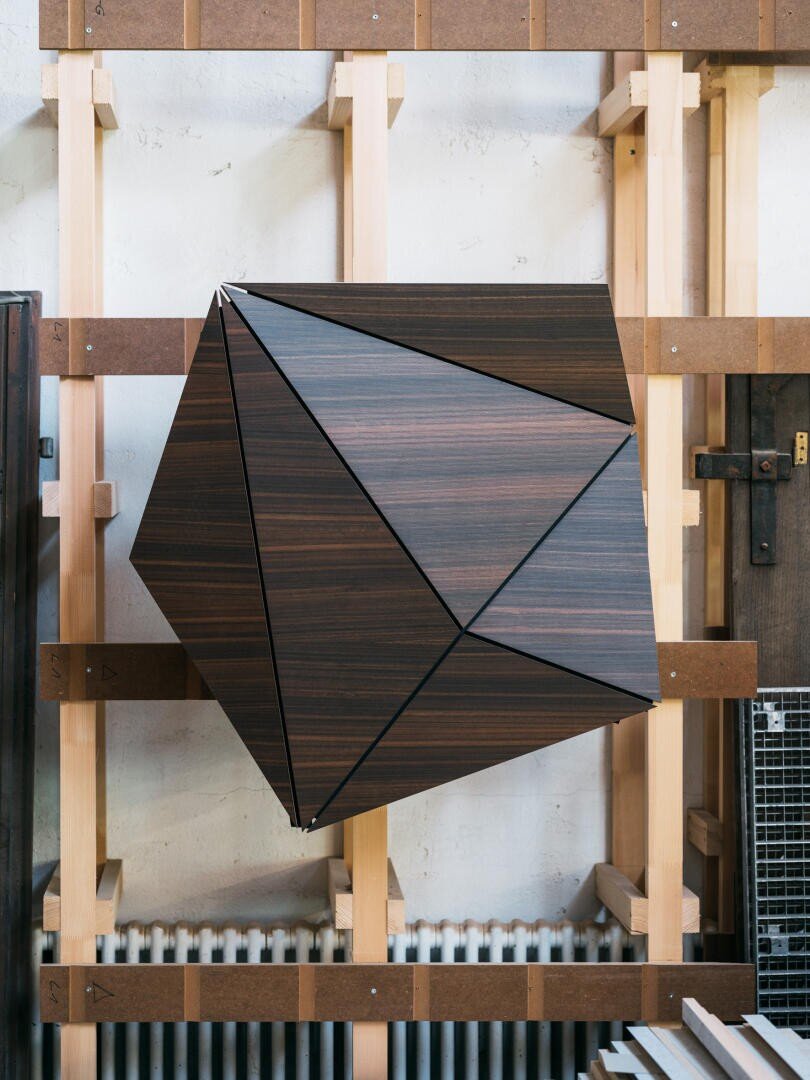 Ein großer geometrischer Holzschrank mit kantigen, facettierten Oberflächen ist auf einem hellen Holzrahmen vor einer weißen Wand montiert. Der Schrank hat eine dunkle Holzoberfläche und schwarze Linien, die seine polygonale Form betonen.