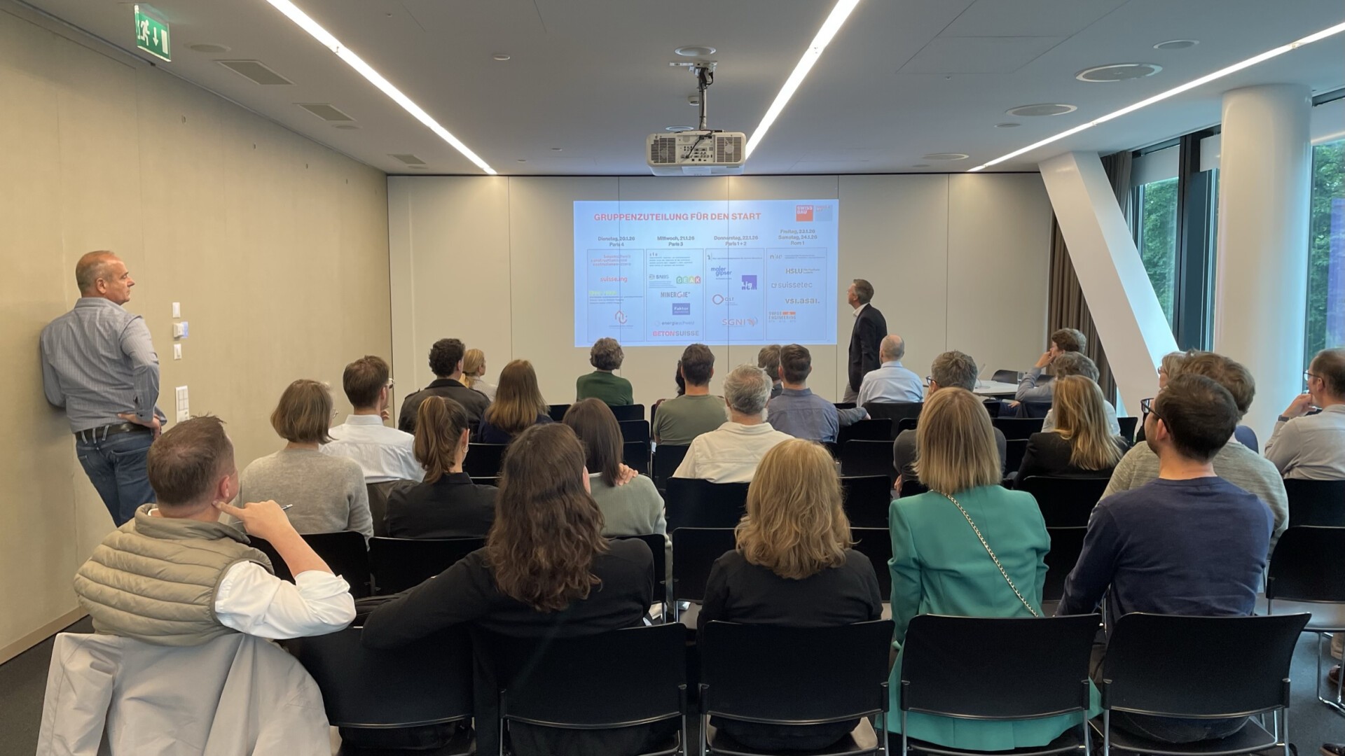 Eine Gruppe von Personen sitzt in einem Konferenzraum und sieht sich eine Präsentation auf einem großen Bildschirm an. Eine Person steht vorne in der Nähe des Bildschirms und hält die Präsentation. Eine andere Person steht seitlich.