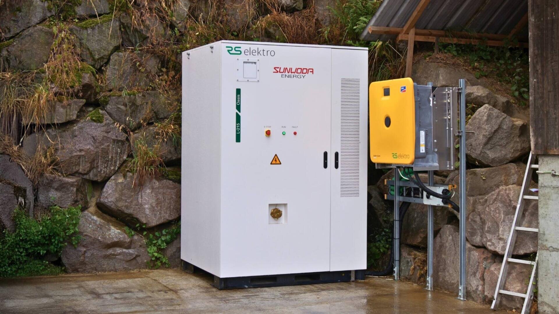 Une grande armoire électrique blanche portant l'inscription "Sunloop Energy" se trouve en plein air sur une surface pavée, près de murs en pierre, à côté d'un appareil jaune plus petit monté sur le mur avec des fils.