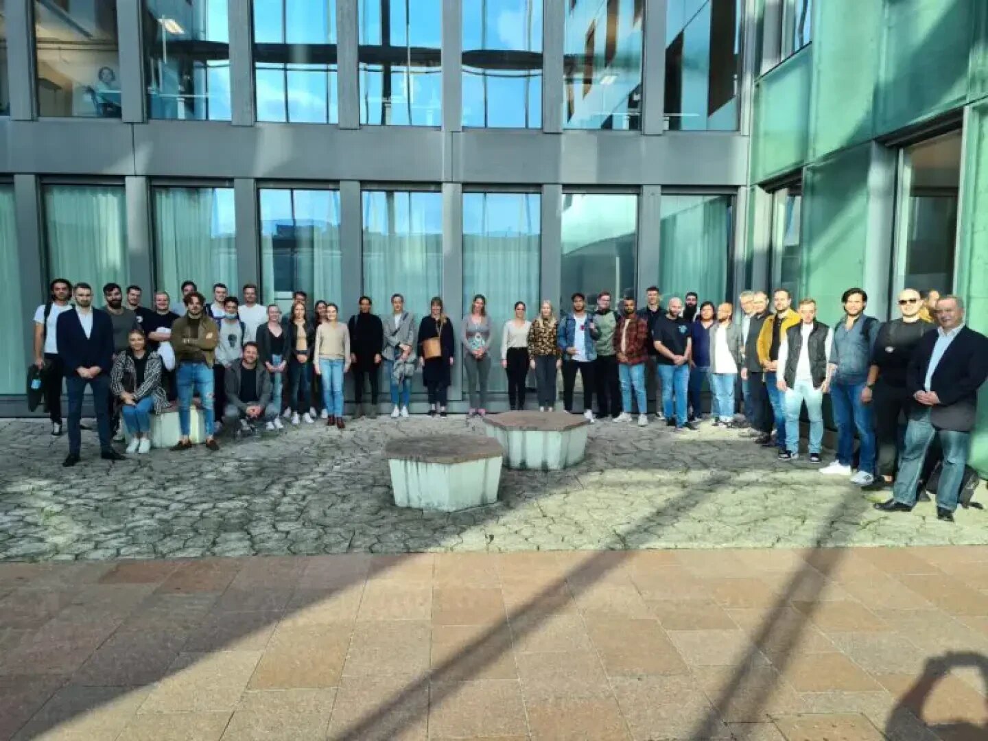 Eine große Gruppe von Menschen steht an einem sonnigen Tag vor einem modernen Glasgebäude und stellt sich für ein Gruppenfoto auf. Die Gruppe ist in einem Halbkreis um zwei runde Steinbänke herum aufgestellt.