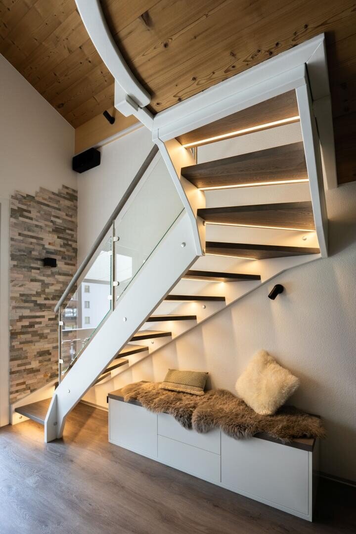 Intérieur moderne avec un escalier en bois avec éclairage intégré, une balustrade en verre et un banc confortable avec des coussins et une couverture en fourrure en dessous, à côté d'un mur avec des accents de pierre et un plafond en bois au-dessus.