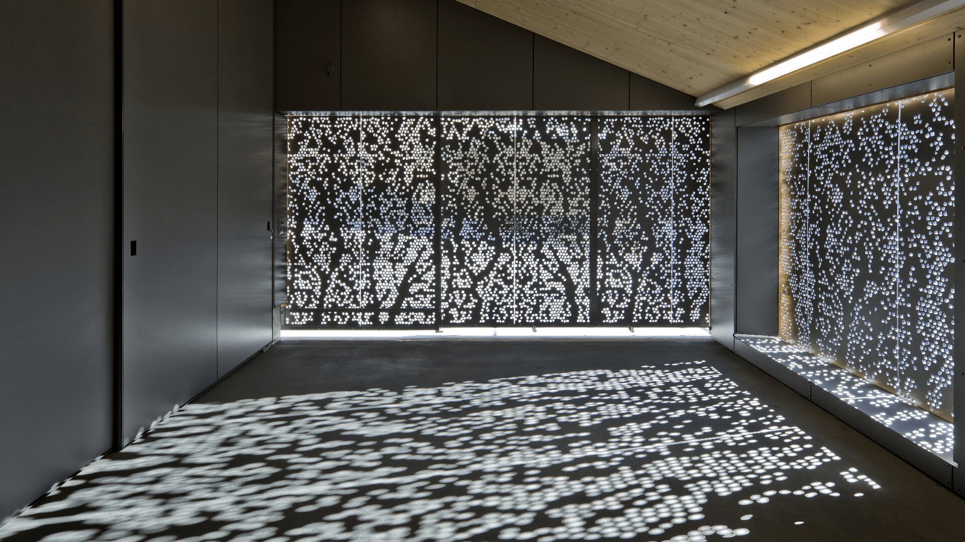 Ein moderner, minimalistischer Raum mit perforierten Metallpaneelen, die es dem Sonnenlicht ermöglichen, auf dem Boden und an den Wänden komplexe Muster aus Pickellicht zu erzeugen. Die Decke ist schräg und aus hellem Holz gefertigt.
