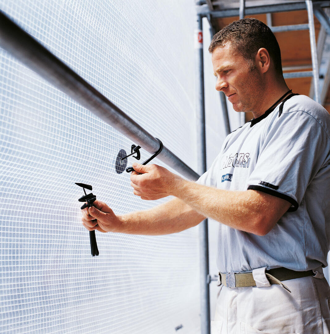 Ein Mann in einem hellgrauen Hemd montiert oder justiert ein kreisförmiges Werkzeug oder eine Halterung an einem Metallmast neben einer mit Maschendraht bedeckten Struktur, möglicherweise auf einer Baustelle.