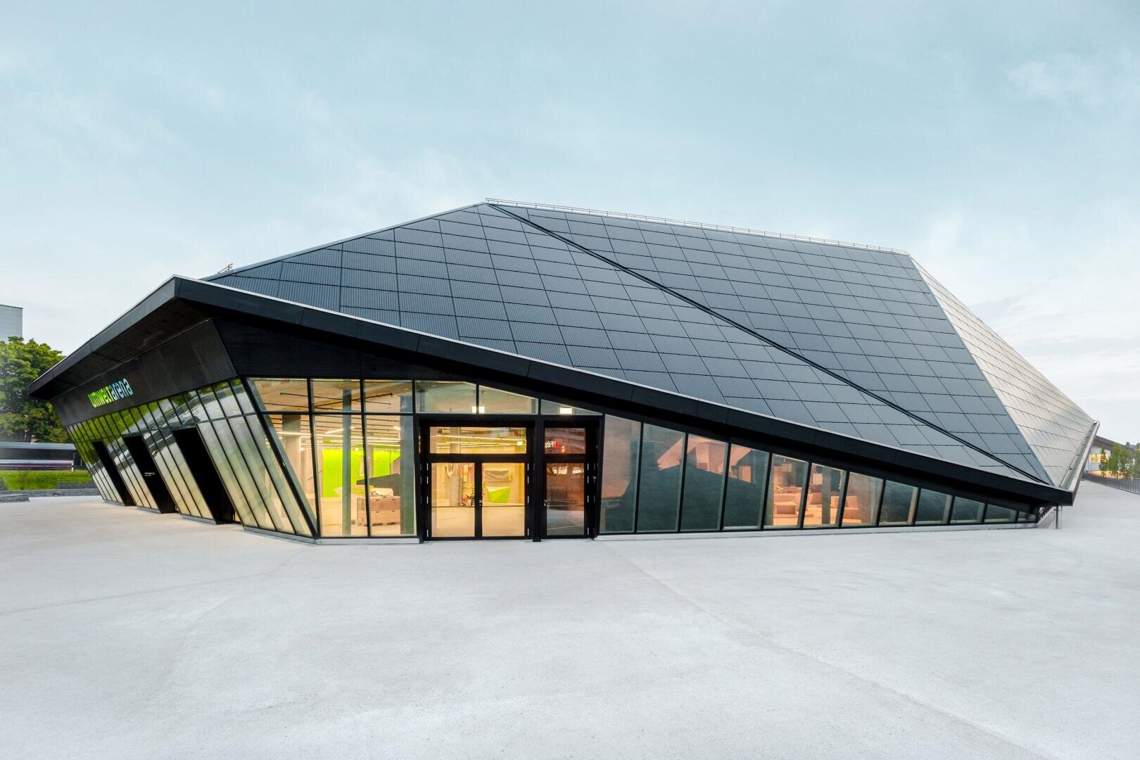Ein modernes Gebäude mit einem kantigen, schrägen Dach und großen Glasfenstern, mit glatten schwarzen Paneelen und einem minimalistischen Design, steht auf einem offenen Betonplatz unter freiem Himmel.