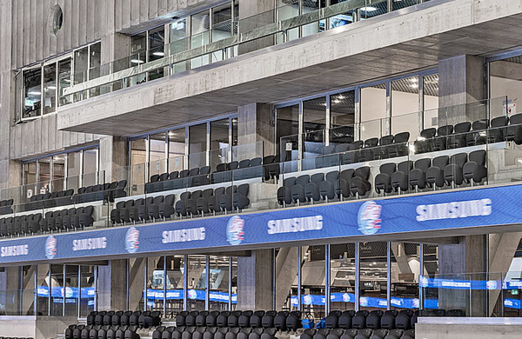Reihen leerer schwarzer Stadionsitze stehen vor einem modernen Betongebäude mit Glasgeländern und großen digitalen Bildschirmen mit dem Samsung-Logo. Der Bereich wirkt sauber und gut beleuchtet.
