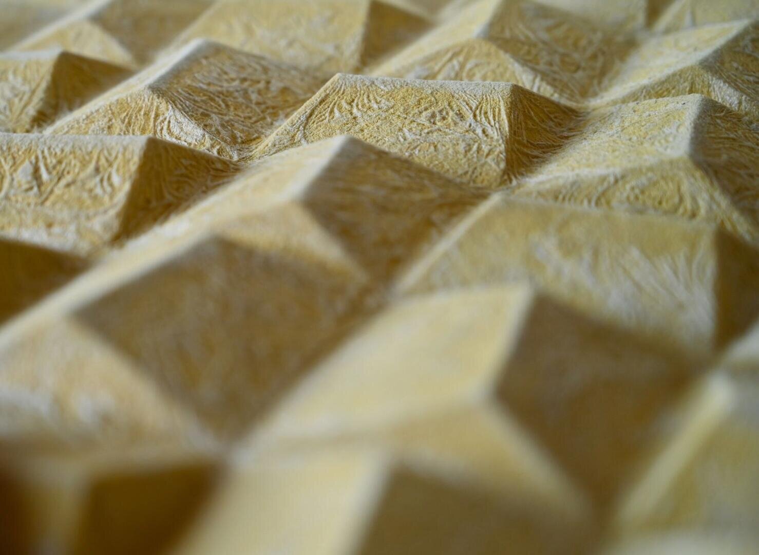 Nahaufnahme einer strukturierten gelben Oberfläche mit sich wiederholenden geometrischen, pyramidenartigen Formen und einer leicht rauen, gemusterten Oberfläche.