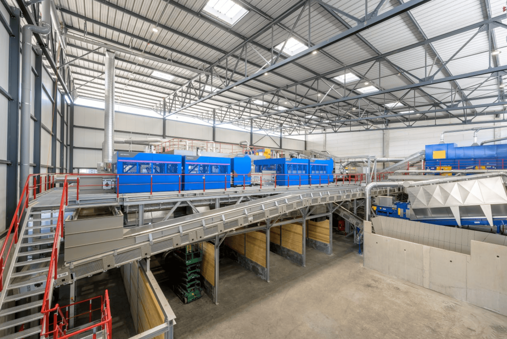 Eine geräumige, moderne Industrieanlage mit Metallstegen, roten Geländern, großen blauen Maschinen und heller Deckenbeleuchtung. Die Szene zeigt einen organisierten Verarbeitungs- oder Fertigungsbereich in einem hohen Lagerhaus.