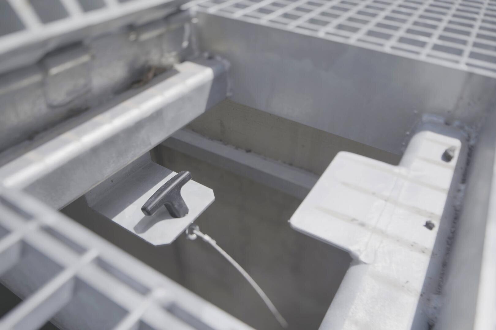 Gros plan sur une structure métallique industrielle avec un fond grillagé, une ouverture rectangulaire et une poignée en forme de T fixée à un câble à l'intérieur de l'ouverture. La couleur générale est gris métallique.