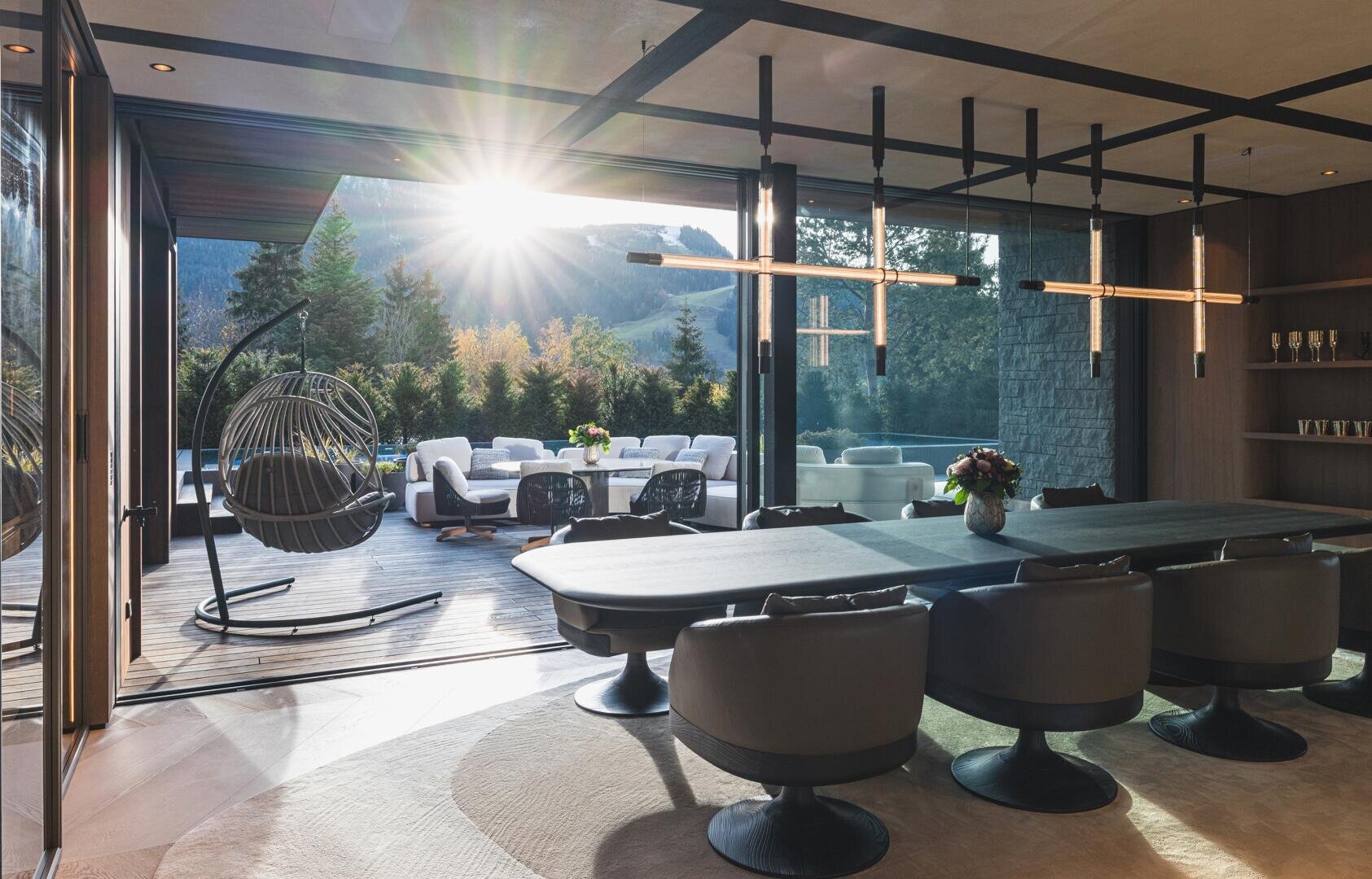 Moderner Essbereich mit schwarzen Stühlen und einem Tisch, der sich zu einer sonnenbeschienenen Außenterrasse mit Terrassenbestuhlung, einem Hängesessel und Blick auf die grüne Landschaft und die fernen Hügel öffnet. Das Sonnenlicht strömt durch große Glastüren.
