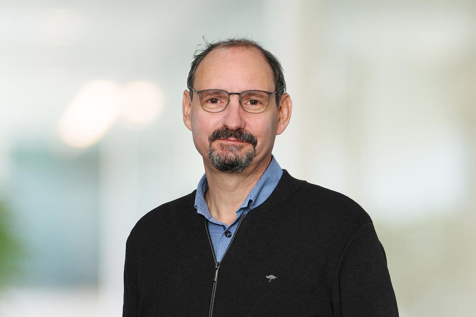 Jörg Kradolfer, ein Gipsermeister mittleren Alters mit kurzen dunklen Haaren, Bart und Brille, der einen schwarzen Pullover mit Reißverschluss über einem blauen Hemd mit Kragen trägt, steht vor einem leicht verschwommenen Innenhintergrund.