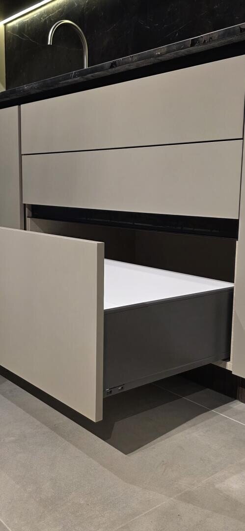 Une cuisine moderne avec des armoires beiges élégantes ; le tiroir inférieur est ouvert, révélant un intérieur blanc spacieux. Le plan de travail est sombre et un robinet est visible à l'arrière-plan. Le sol est en carrelage gris.