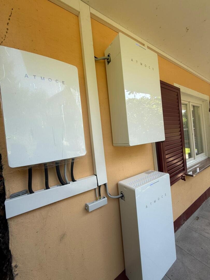 Drei weiße Elektrogeräte mit der Aufschrift ATMOCE sind an einer gelben Außenwand in der Nähe eines Fensters mit braunen Fensterläden angebracht. Schwarze Kabel führen von der linken Einheit zu den beiden gestapelten Kästen auf der rechten Seite.