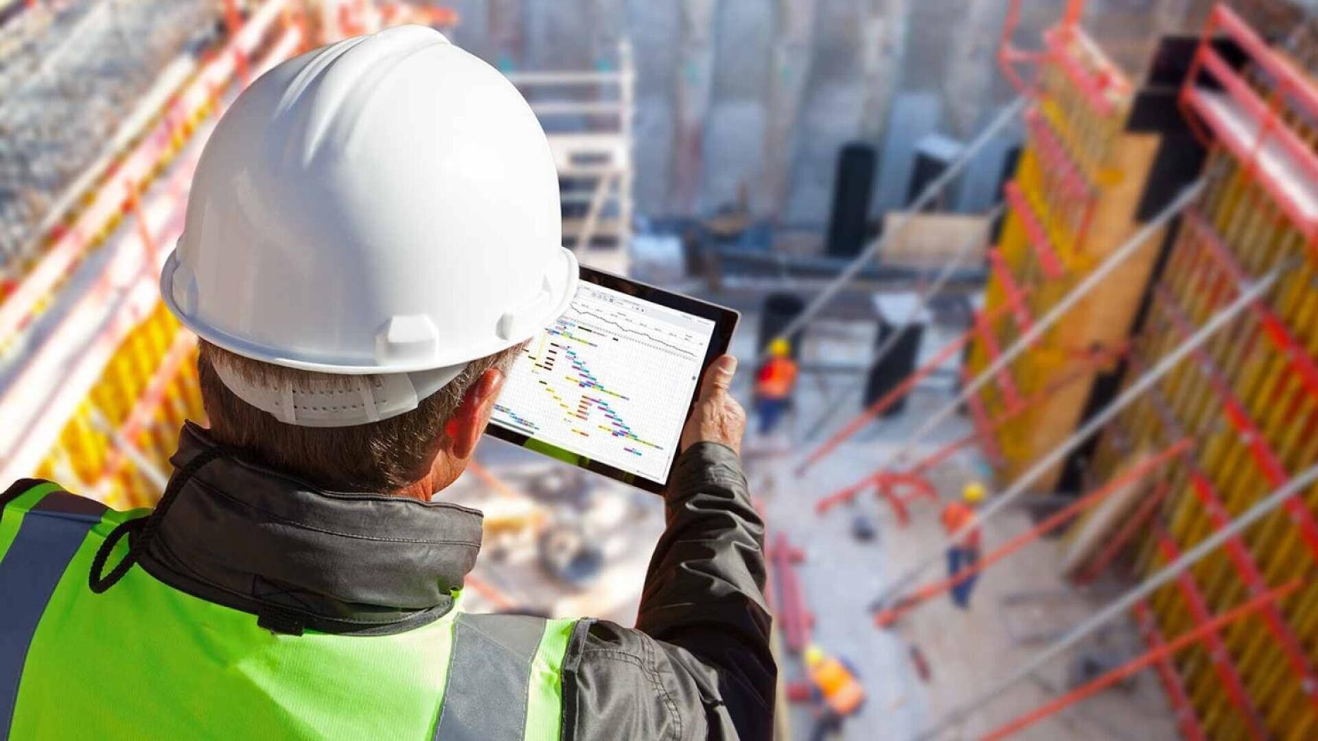 Un ouvrier du bâtiment portant un casque blanc et un gilet de sécurité jaune vérifie une tablette sur laquelle est affiché un plan de projet, sur un chantier animé avec des échafaudages et des ouvriers en arrière-plan.