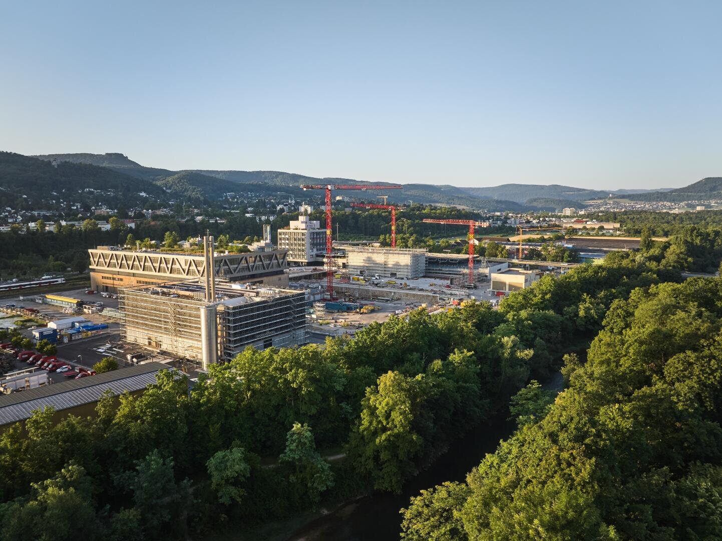 Luftaufnahme einer Großbaustelle mit mehreren Kränen und modernen Gebäuden, umgeben von grünen Bäumen und Hügeln unter einem klaren Himmel.