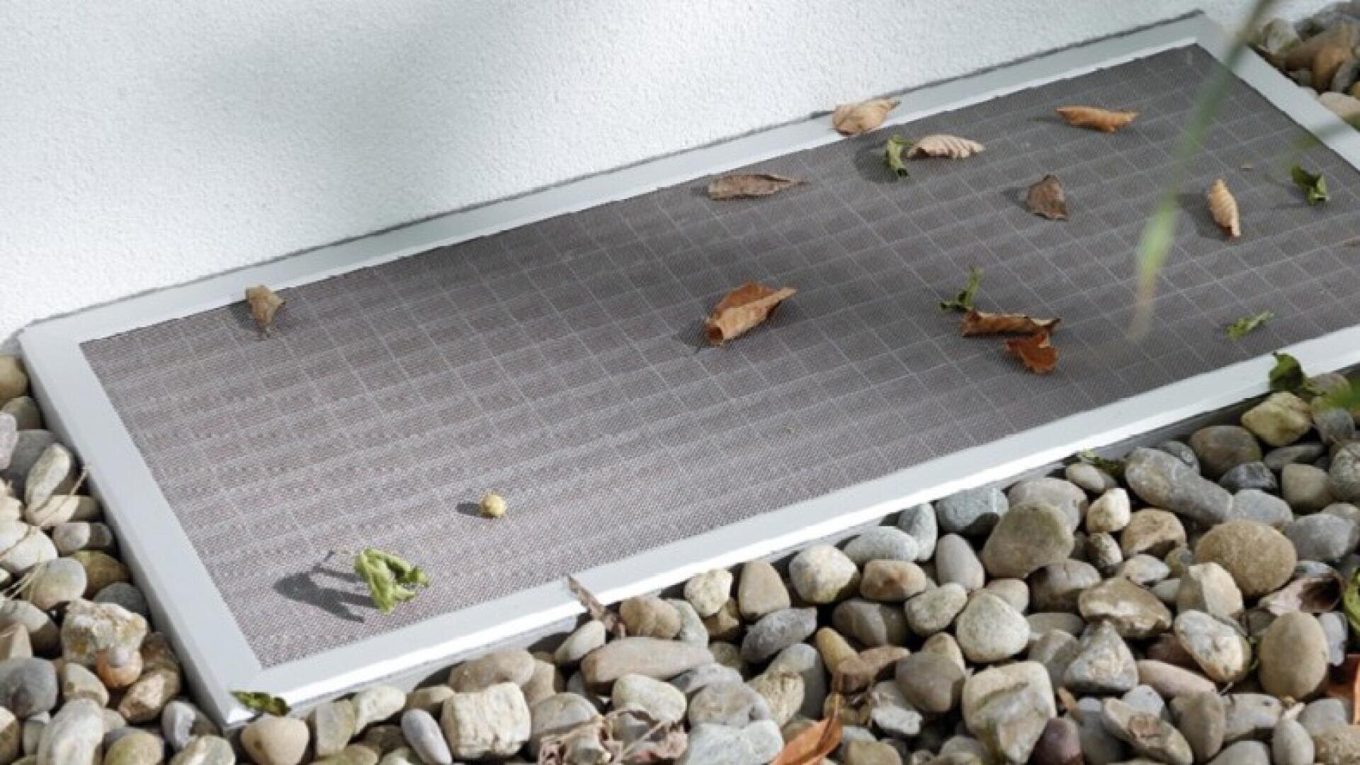 Ein metallenes Lüftungsgitter mit einem Maschensieb ist in den Boden eingelassen, umgeben von kleinen Steinen und verstreuten trockenen Blättern. Hinter dem Gitter ist eine weiße Wand zu sehen.