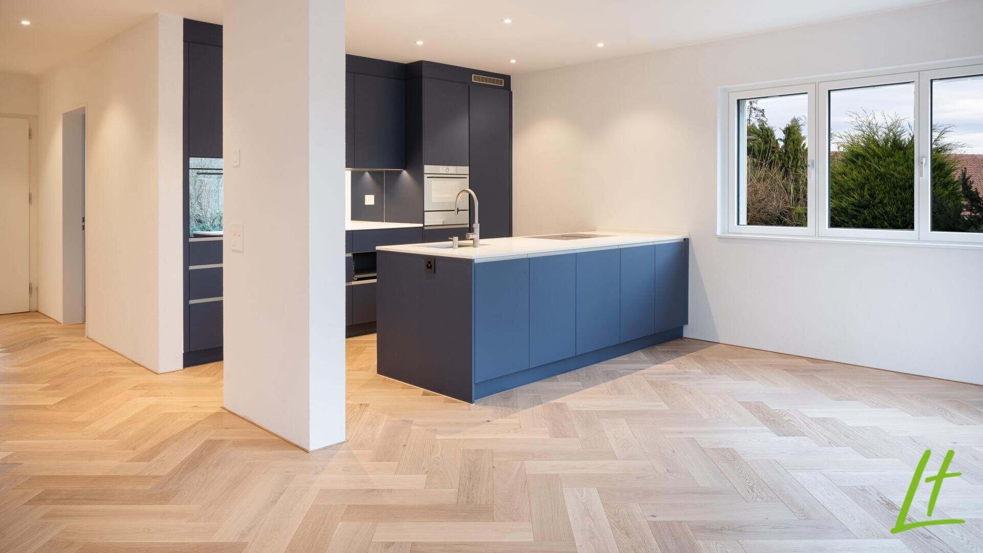 Moderne Küche mit blauen Schränken, weißen Arbeitsplatten, Einbaugeräten und einer zentralen Insel. Der Raum ist mit hellem Fischgrätparkett, weißen Wänden und großen Fenstern ausgestattet, die viel natürliches Licht hereinlassen.