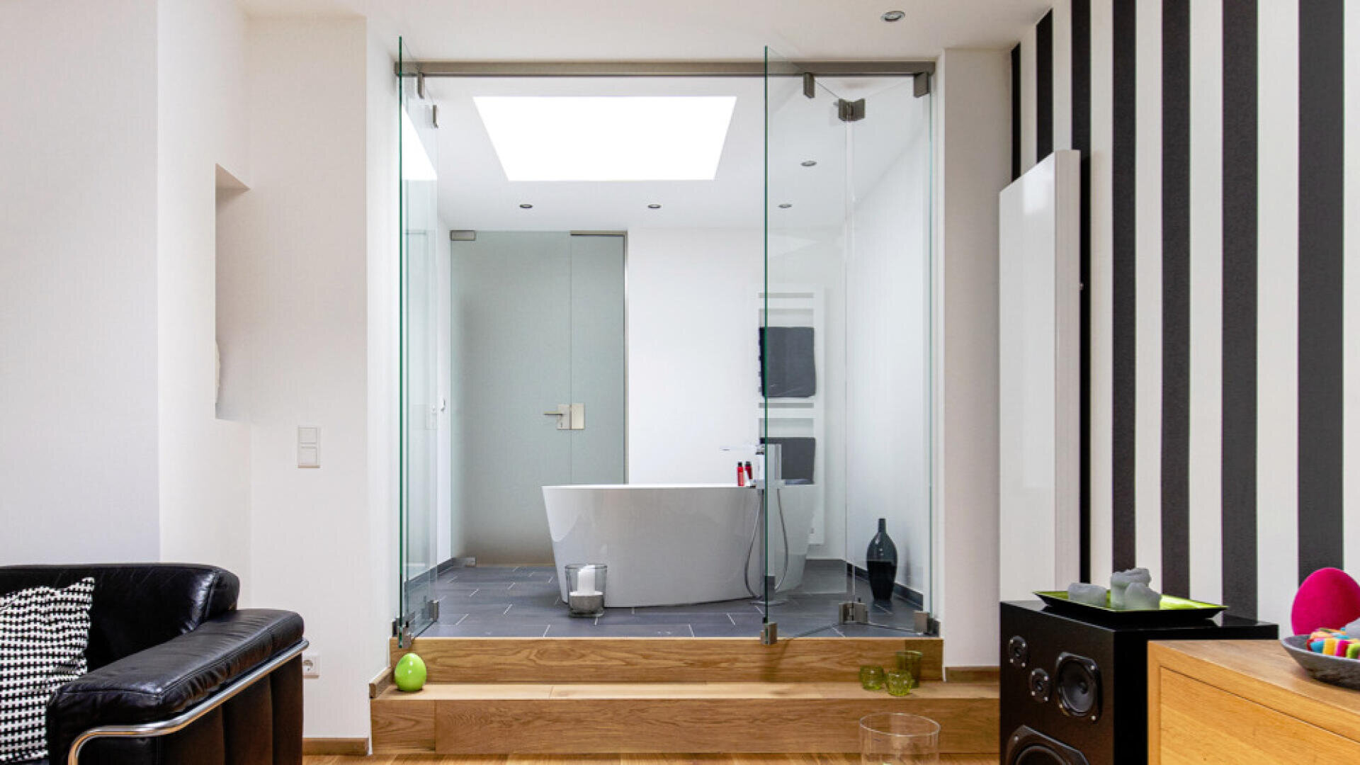 Ein modernes Badezimmer mit freistehender Badewanne und Glaswänden ist von einem Wohnbereich mit Holzboden, schwarzem Ledersessel, gestreifter Wand und farbenfrohen Dekorationsgegenständen aus zu sehen. Der Raum ist hell mit natürlichem Licht.
