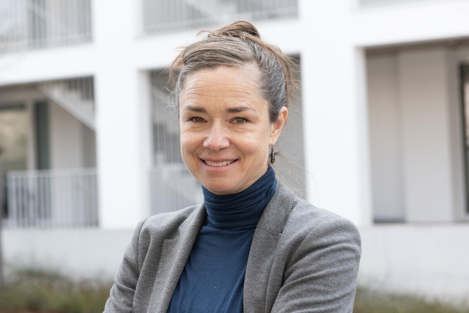 Eine Frau mit braunen Haaren in einem Dutt, die einen grauen Blazer und einen blauen Rollkragenpullover trägt, lächelt, während sie im Freien vor einem weißen Gebäude mit Geländer und Treppenaufgang steht.