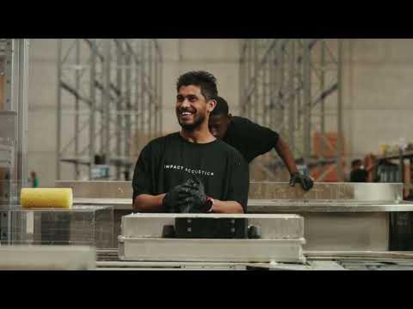 Ein Mann mit schwarzen Handschuhen und einem schwarzen T-Shirt lächelt, während er an einer Werkbank in einem Industrielager steht, mit Metallregalen und einem anderen Arbeiter im Hintergrund.