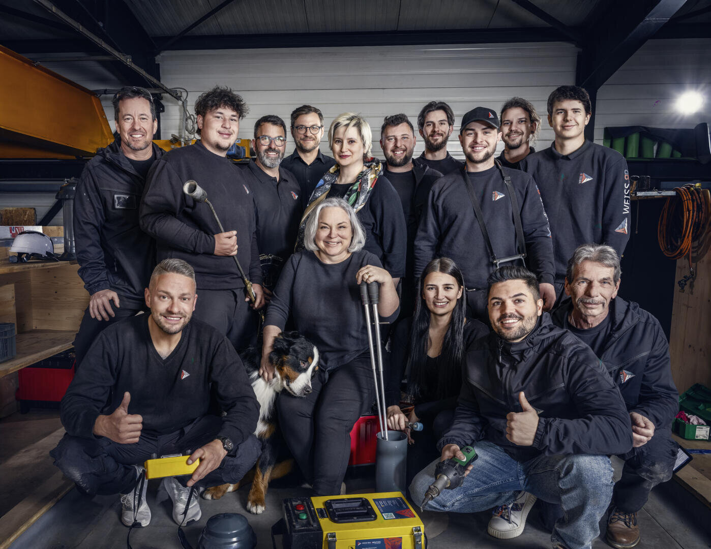 Un groupe de quinze personnes, dont certaines tiennent des outils à la main, posent et sourient ensemble dans un atelier. Une femme au centre est assise avec un chien. En arrière-plan, on voit des étagères, des outils et des machines.