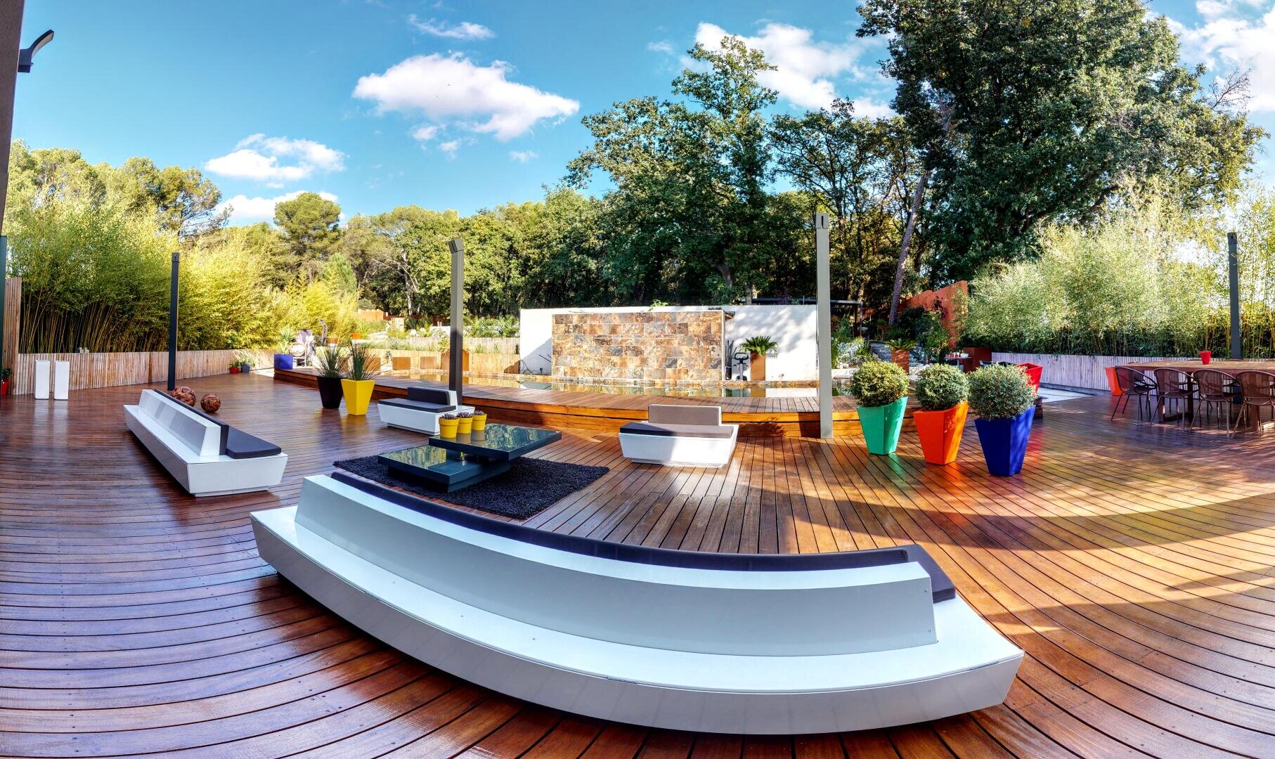 Un patio extérieur spacieux avec des bancs blancs incurvés, une table basse en verre, des plantes en pot colorées, une terrasse en bois et des arbres en arrière-plan sous un ciel partiellement nuageux.