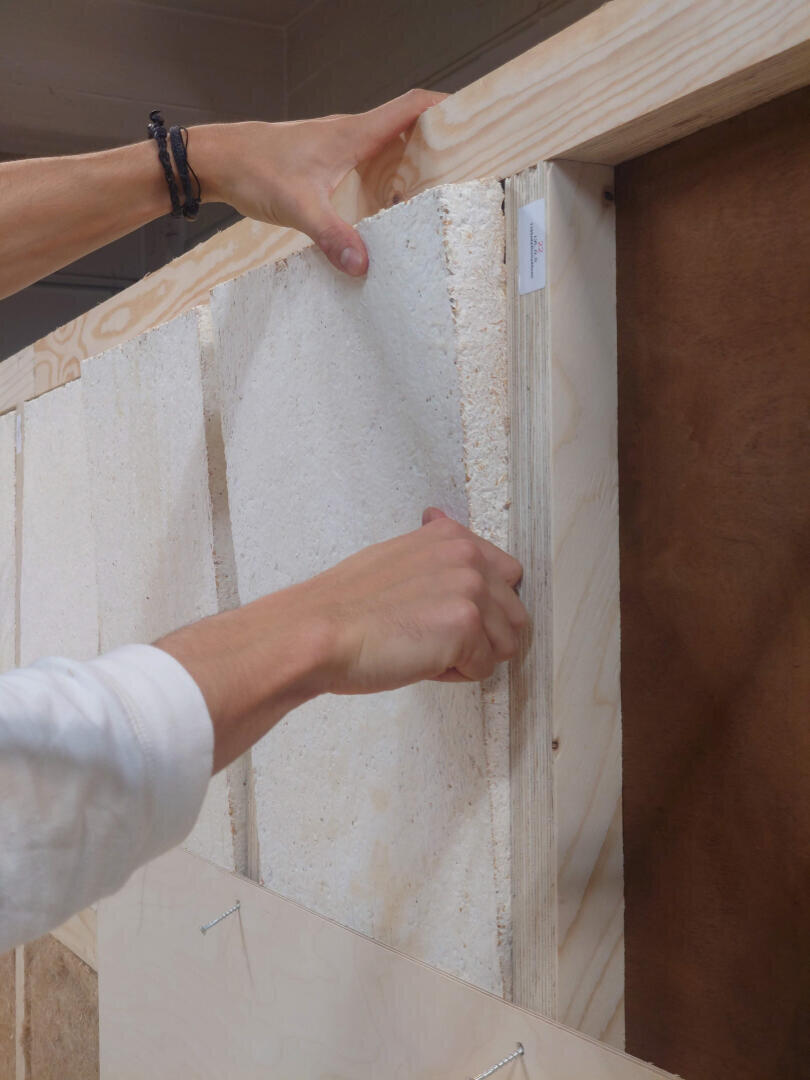 Eine Person bringt rechteckige weiße Dämmplatten in einem Holzrahmen an und drückt eine Platte mit beiden Händen fest. Die Szene scheint Teil eines Bau- oder Renovierungsprojekts zu sein.