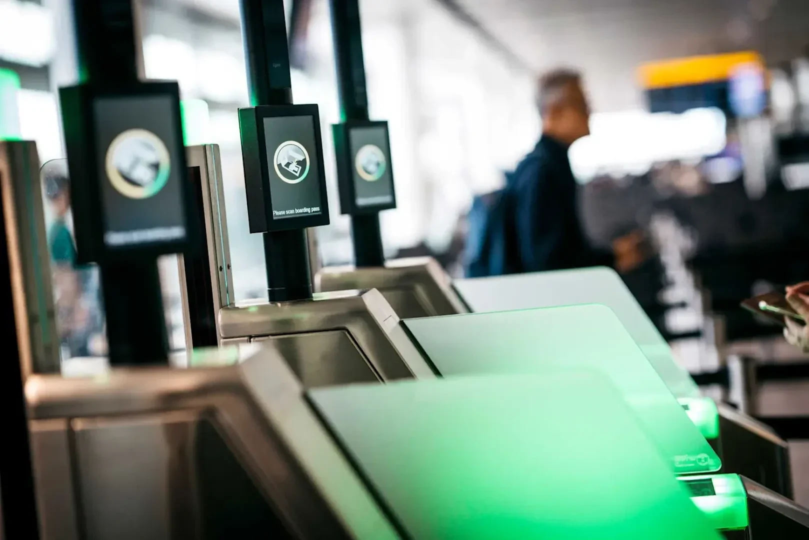 Eine Reihe automatischer Passkontrollschleusen mit grünen Lichtern auf einem Flughafen mit digitalen Bildschirmen, auf denen ID-Symbole angezeigt werden; im Hintergrund ist eine Person zu sehen.