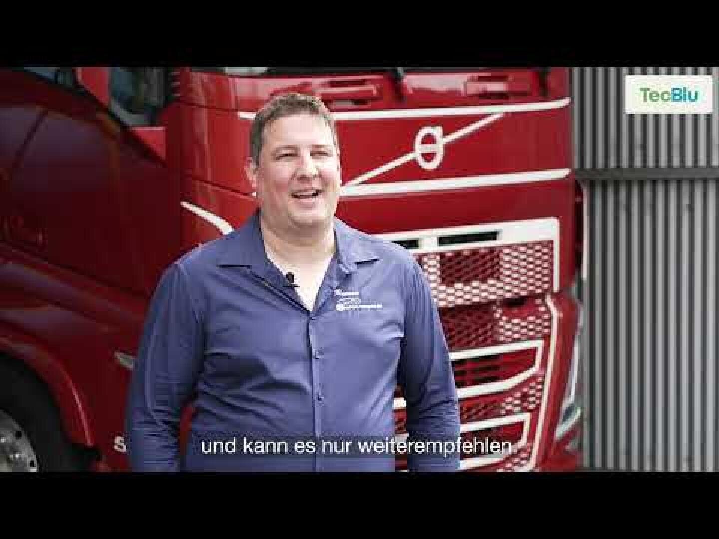 Ein lächelnder Mann in einem blauen Hemd steht vor einem roten Volvo-Lkw. Auf dem Bild erscheinen deutsche Untertitel und ein TecBlu-Logo.