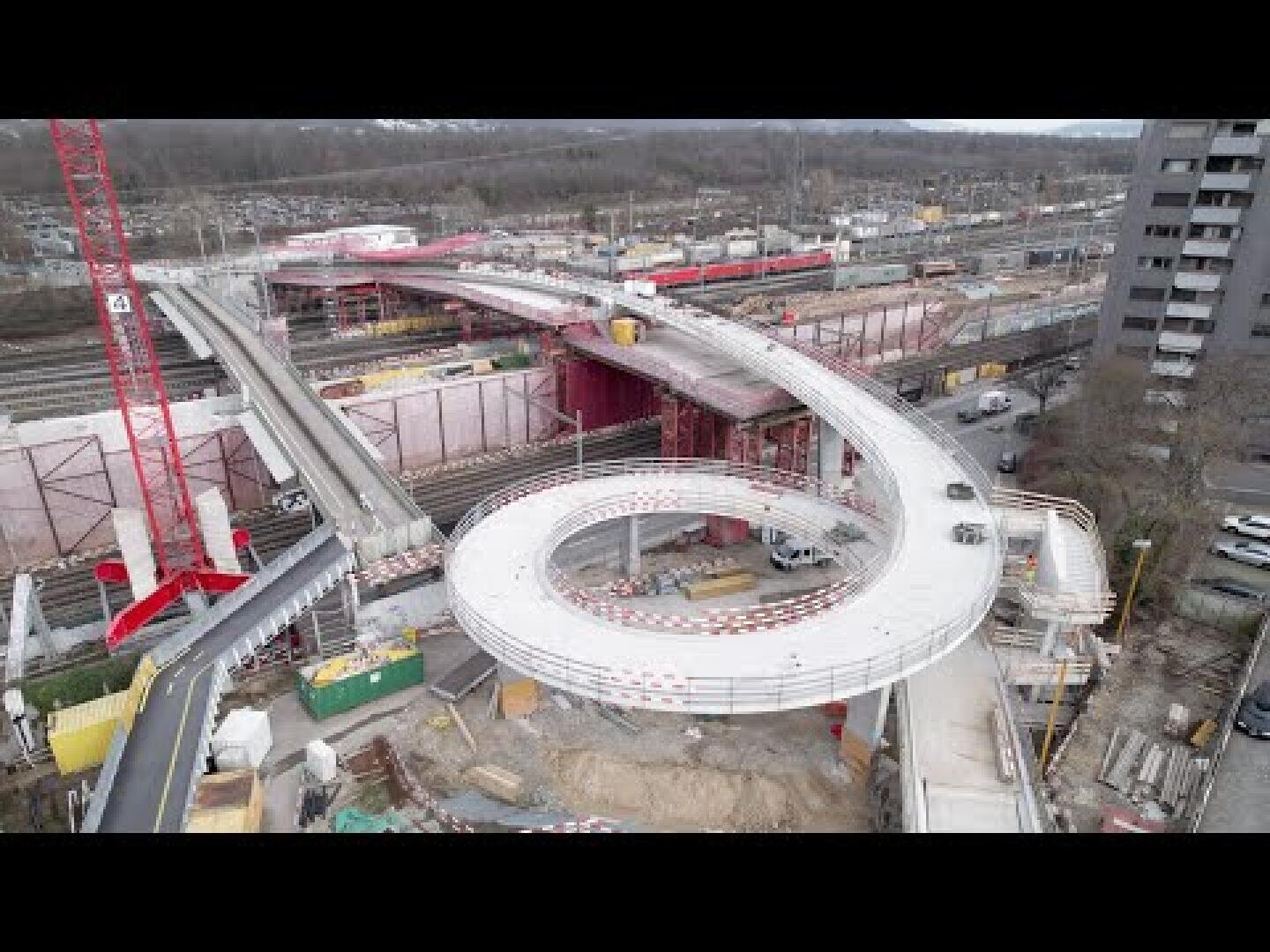 Luftaufnahme einer Großbaustelle mit einer weißen spiralförmigen Fußgänger- oder Fahrradbrücke im Bau, umgeben von Kränen, Gebäuden und verschiedenen Baumaterialien.