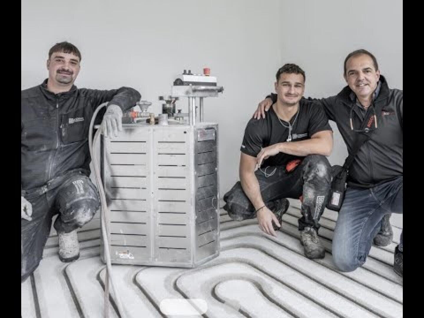 Drei Männer in Arbeitskleidung knien auf einem Boden mit freiliegenden Fußbodenheizungsrohren und lächeln in die Kamera. Sie posieren neben einer Metallmaschine, die für die Installation oder Prüfung des Heizungssystems verwendet wird.