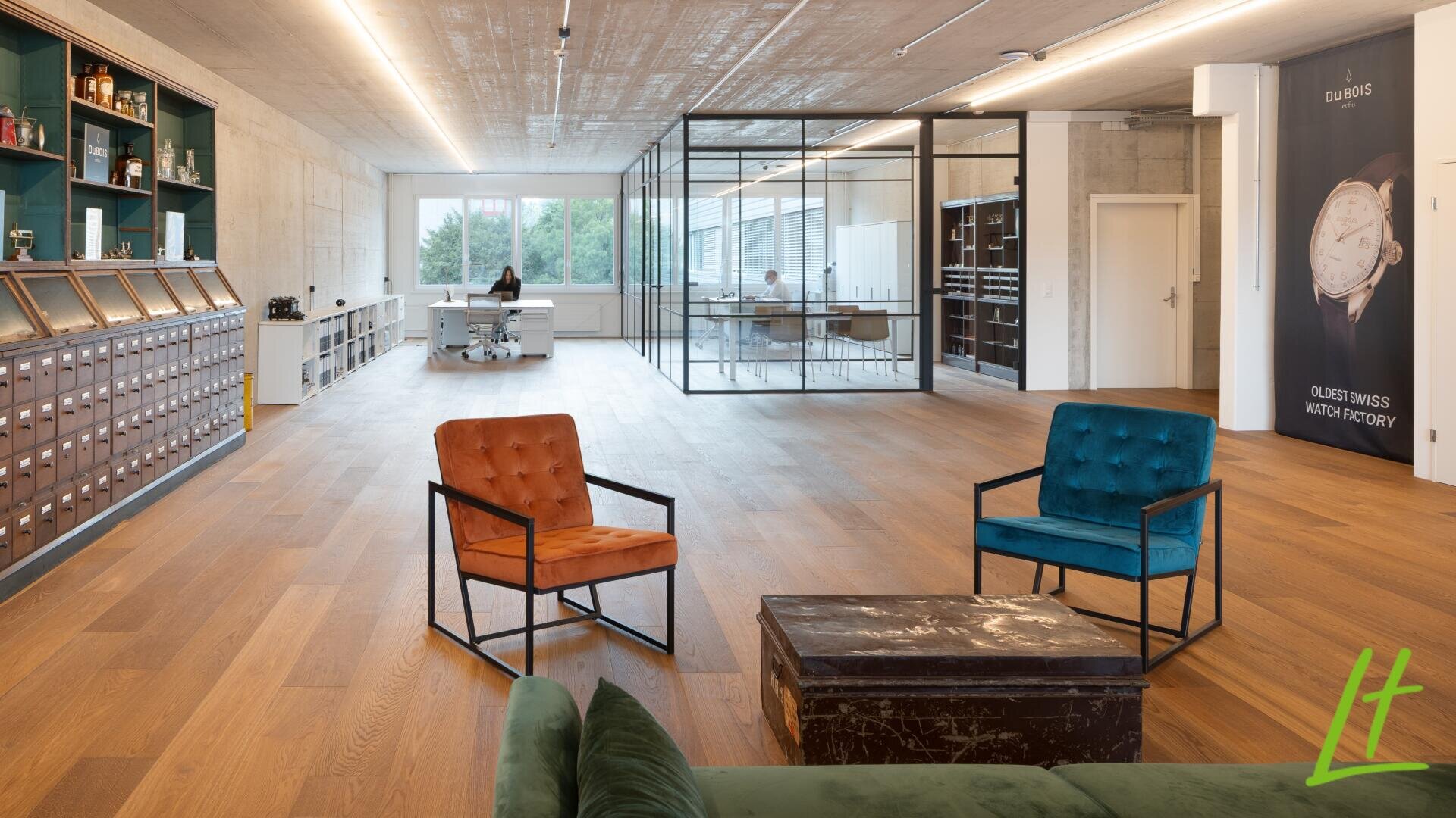 Modernes Großraumbüro mit Holzboden, Sichtbetondecke, Loungesesseln, grünem Sofa, alten Schränken und einem gläsernen Besprechungsraum. Zwei Personen arbeiten an Schreibtischen in der Nähe großer Fenster im Hintergrund.
