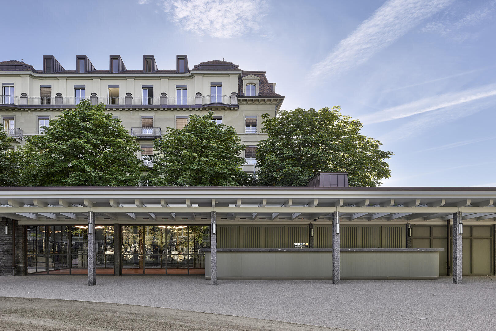 Ein moderner Glas- und Metallpavillon mit Flachdach steht vor einem hohen, historischen Gebäude mit Balkonen. Bäume verdecken teilweise die ältere Fassade. Der Himmel über dem Gebäude ist klar mit Wolkenschlieren.