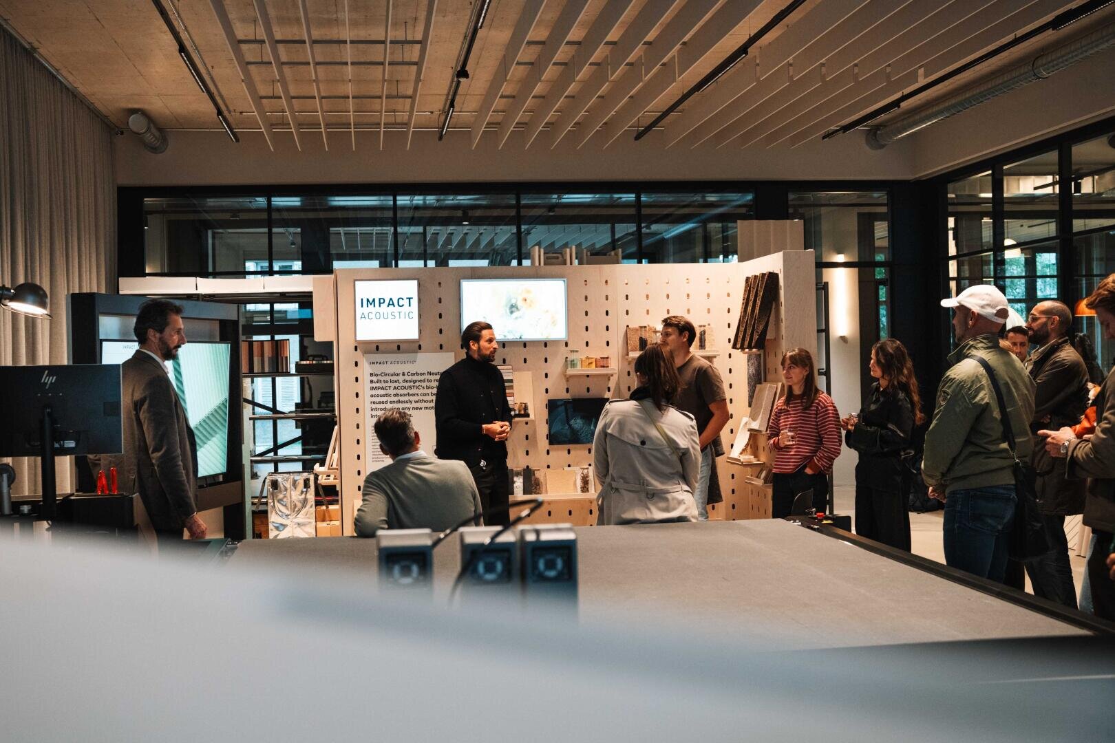 Eine Gruppe von Personen hört einem Vortragenden in einem modernen Büroraum mit großen Fenstern zu. Hinter dem Vortragenden befindet sich eine Ausstellungswand mit der Aufschrift "Impact Acoustic". Einige Zuhörer sitzen, andere stehen.