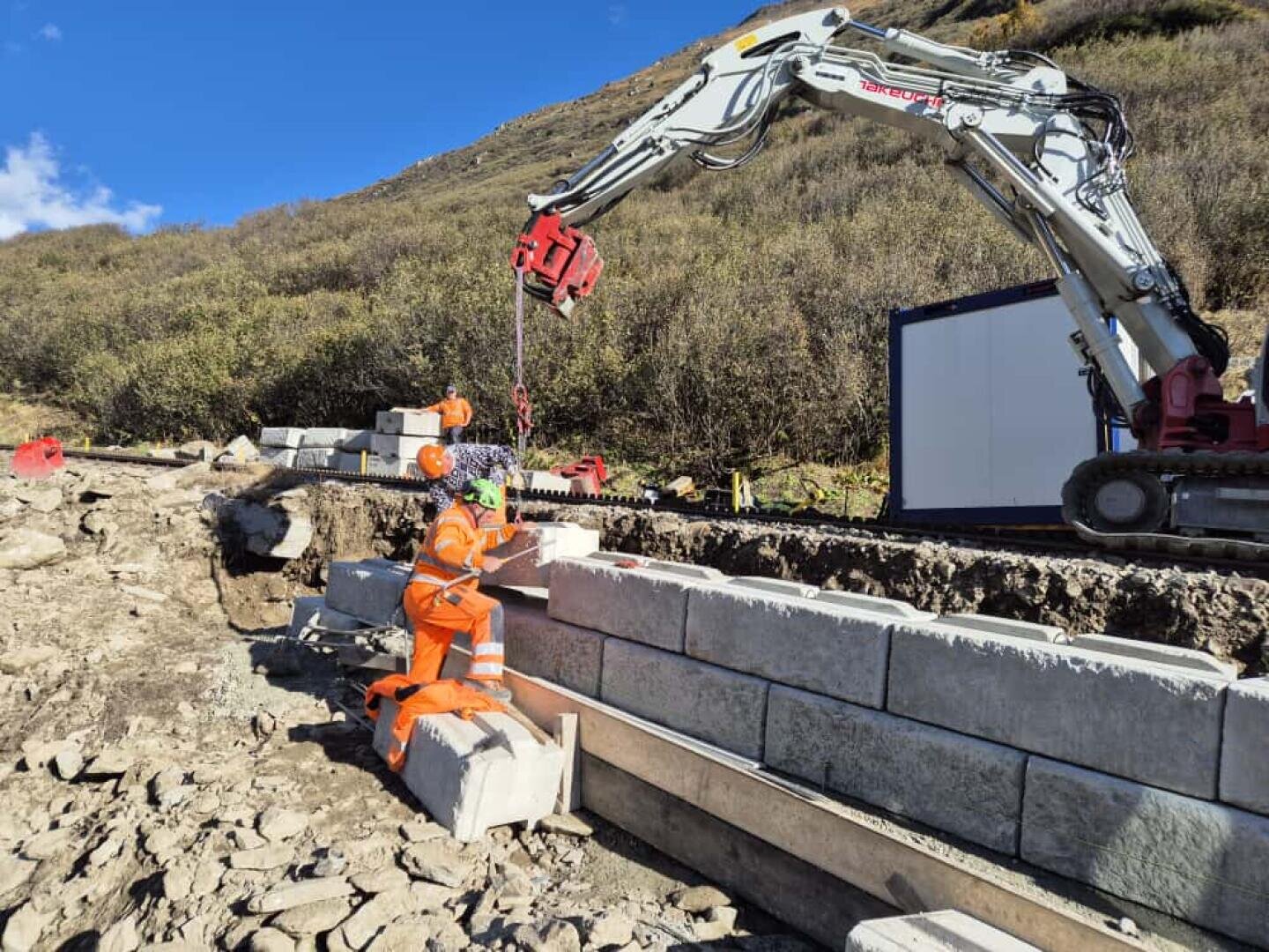 Zwei Bauarbeiter in orangefarbener Schutzkleidung und mit Helmen bringen große Betonblöcke mit Hilfe eines Baggers auf einer Baustelle im Freien inmitten von Erde und Hügeln in Position.